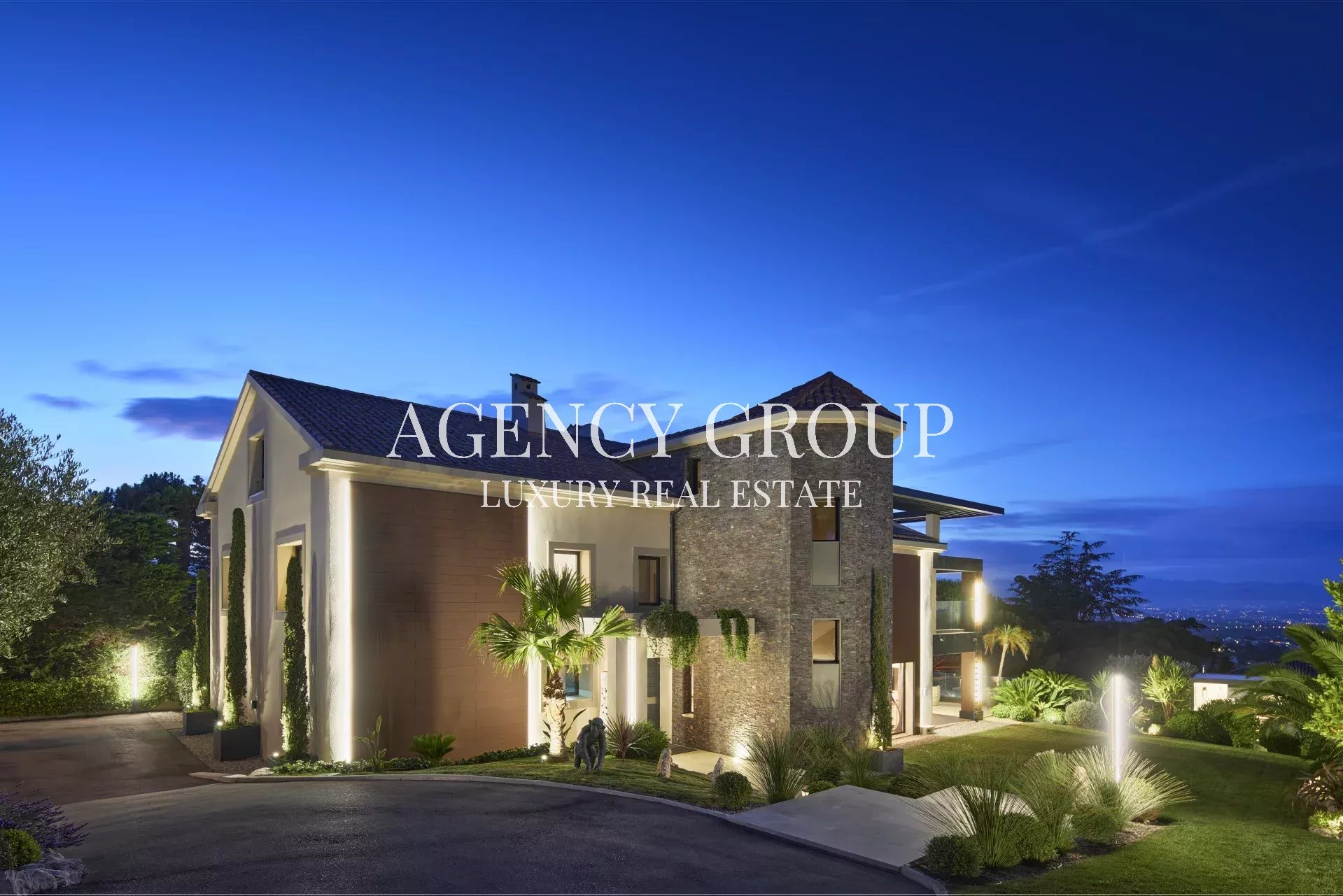 Twilight exterior of a modern luxury house with landscaped grounds and a large 'Agency Group Luxury Real Estate' logo across the facade.