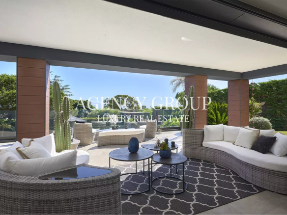 Luxury outdoor living space on a covered patio with a curved wicker sofa, round coffee tables, and a glass railing overlooking a green garden.