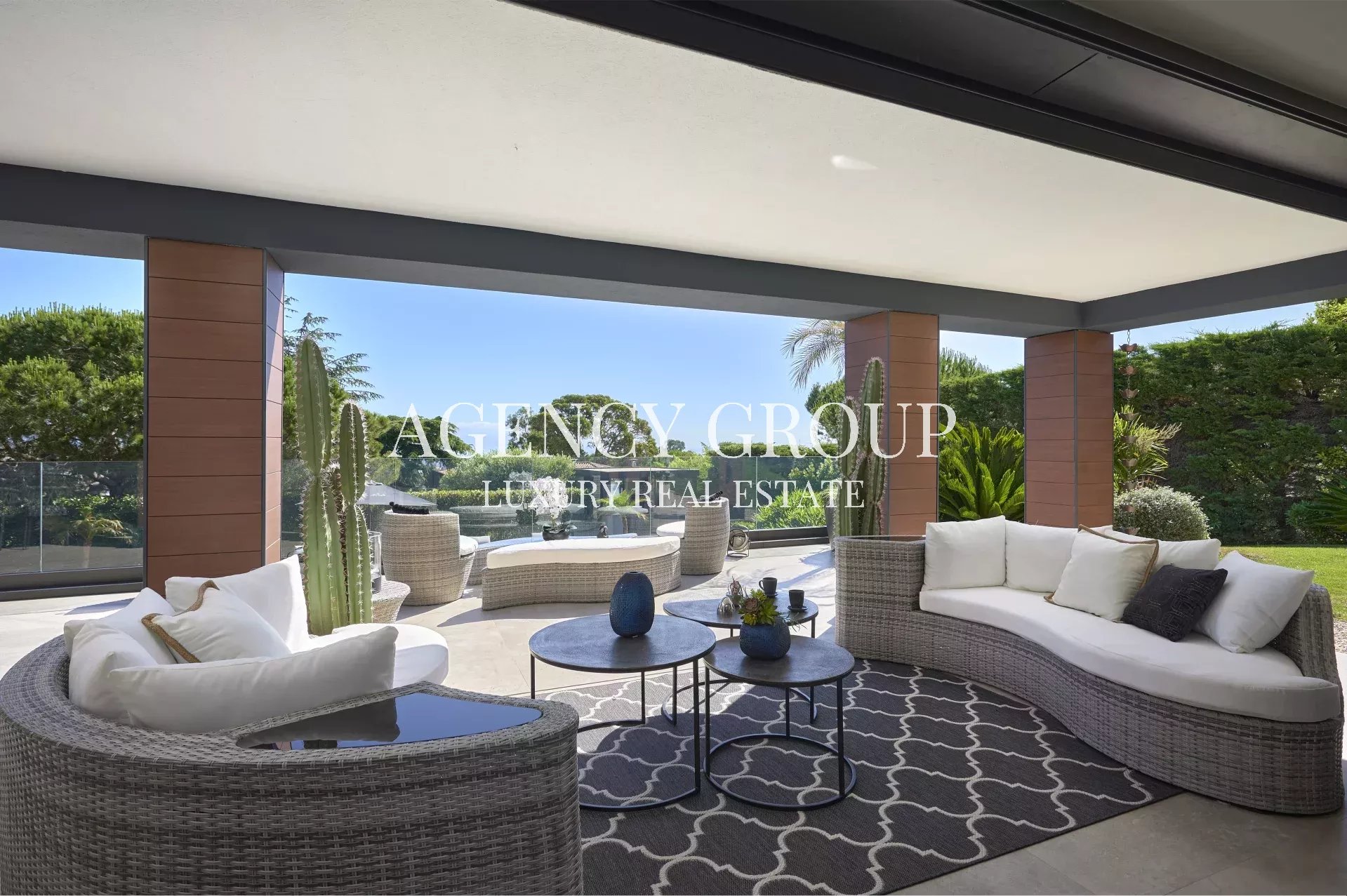Luxury outdoor living space on a covered patio with a curved wicker sofa, round coffee tables, and a glass railing overlooking a green garden.