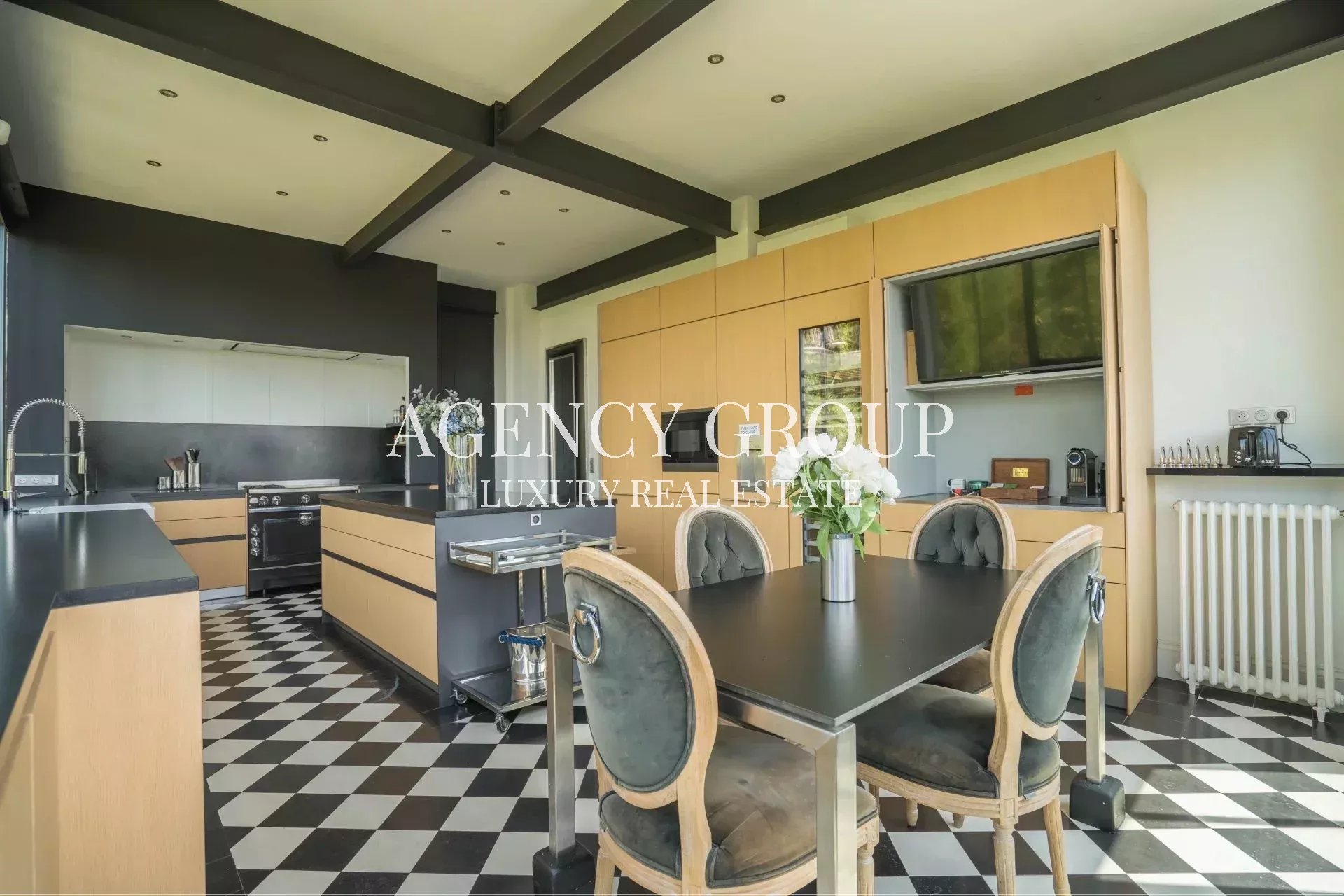Open-plan kitchen with light wood cabinets, black countertops, built-in oven, and a dining table with four upholstered chairs on a black-and-white checkered floor.