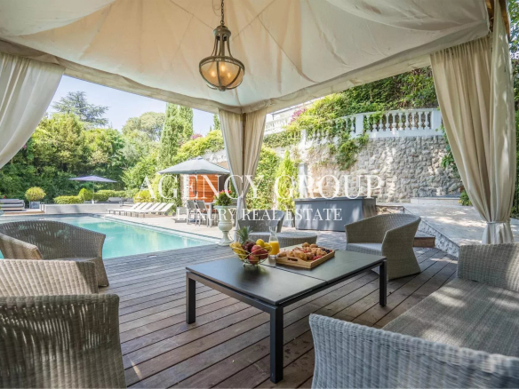 Outdoor poolside lounge under a white canopy with wicker chairs and a coffee table holding fruit and pastries, surrounded by greenery and a stone wall.