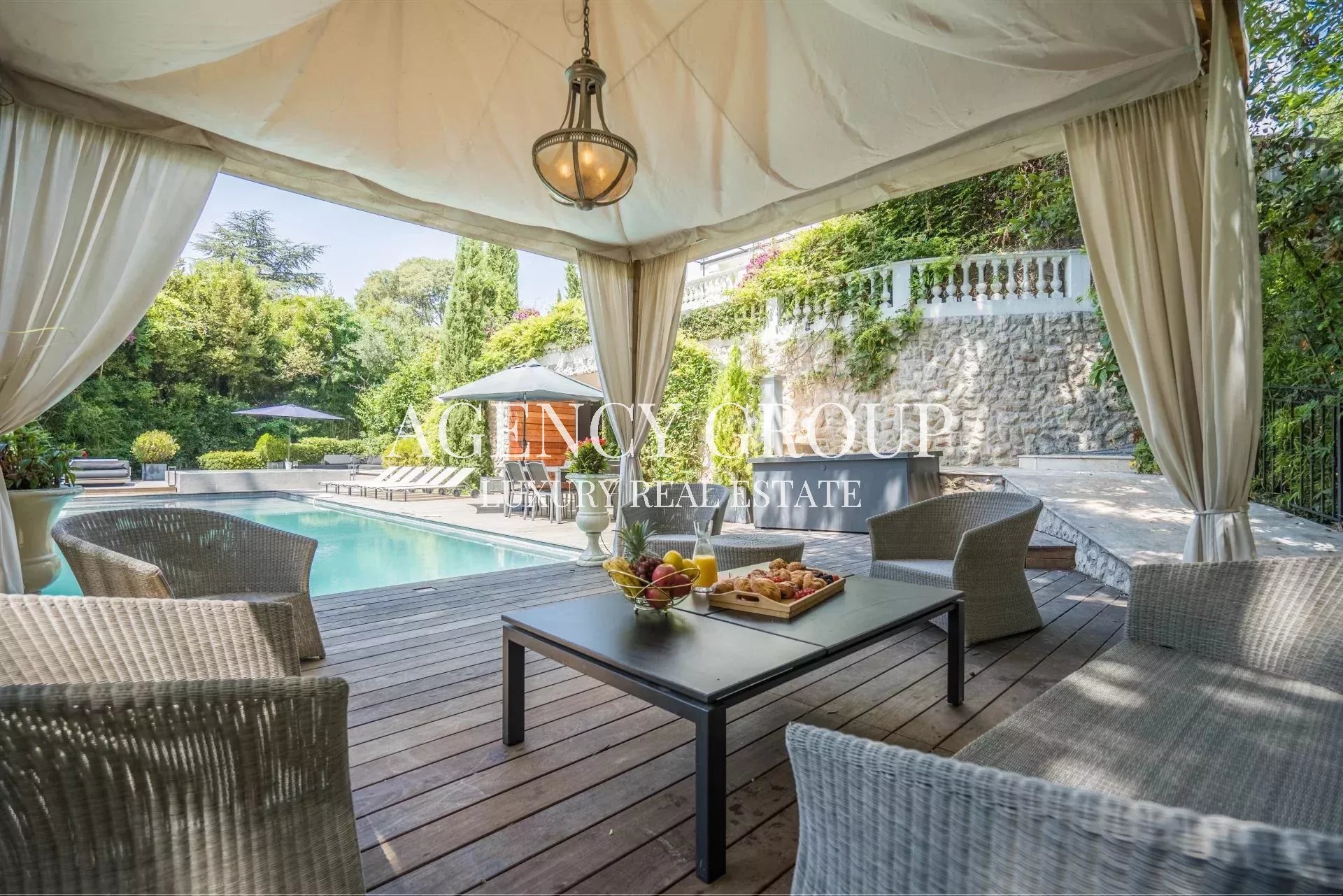 Outdoor poolside lounge under a white canopy with wicker chairs and a coffee table holding fruit and pastries, surrounded by greenery and a stone wall.