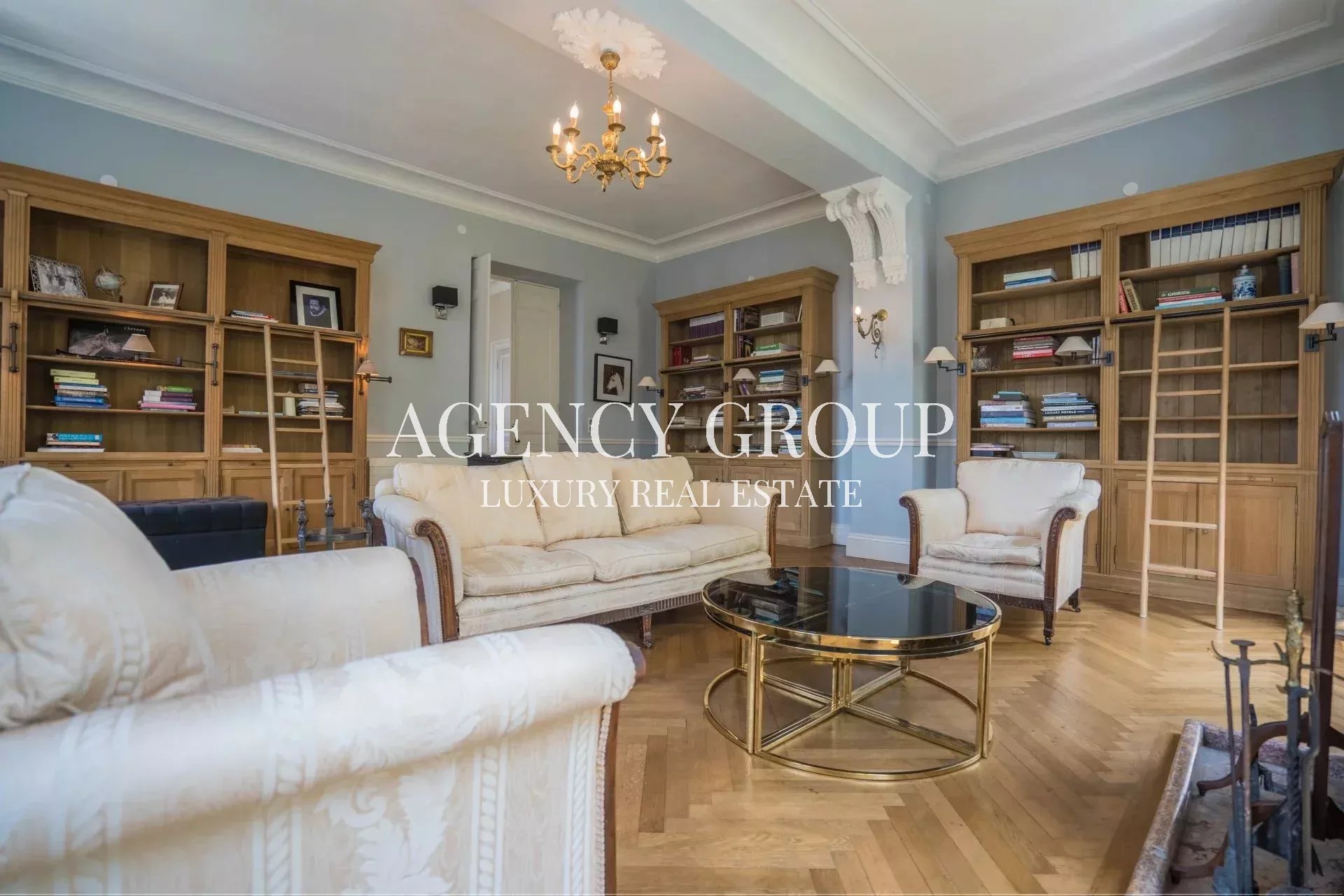 Elegant living room in a luxury home: blue walls, cream sofas, wooden bookcases with ladders, and a gold glass coffee table under a chandelier.