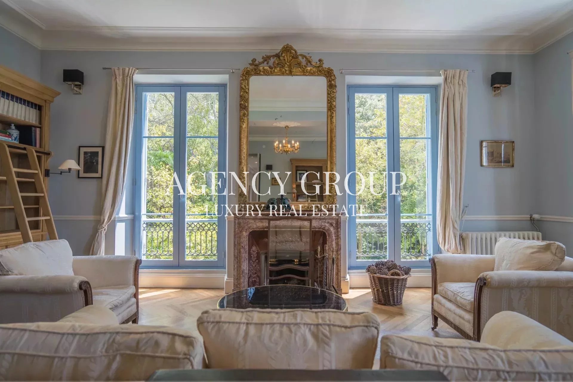 Elegant living room with blue-trimmed windows, cream sofas, and a gold ornate mirror above a marble fireplace.