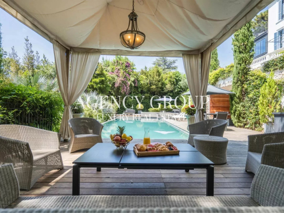 Outdoor cabana-style seating area with cream curtains, a central coffee table, and a view of a rectangular pool surrounded by greenery.