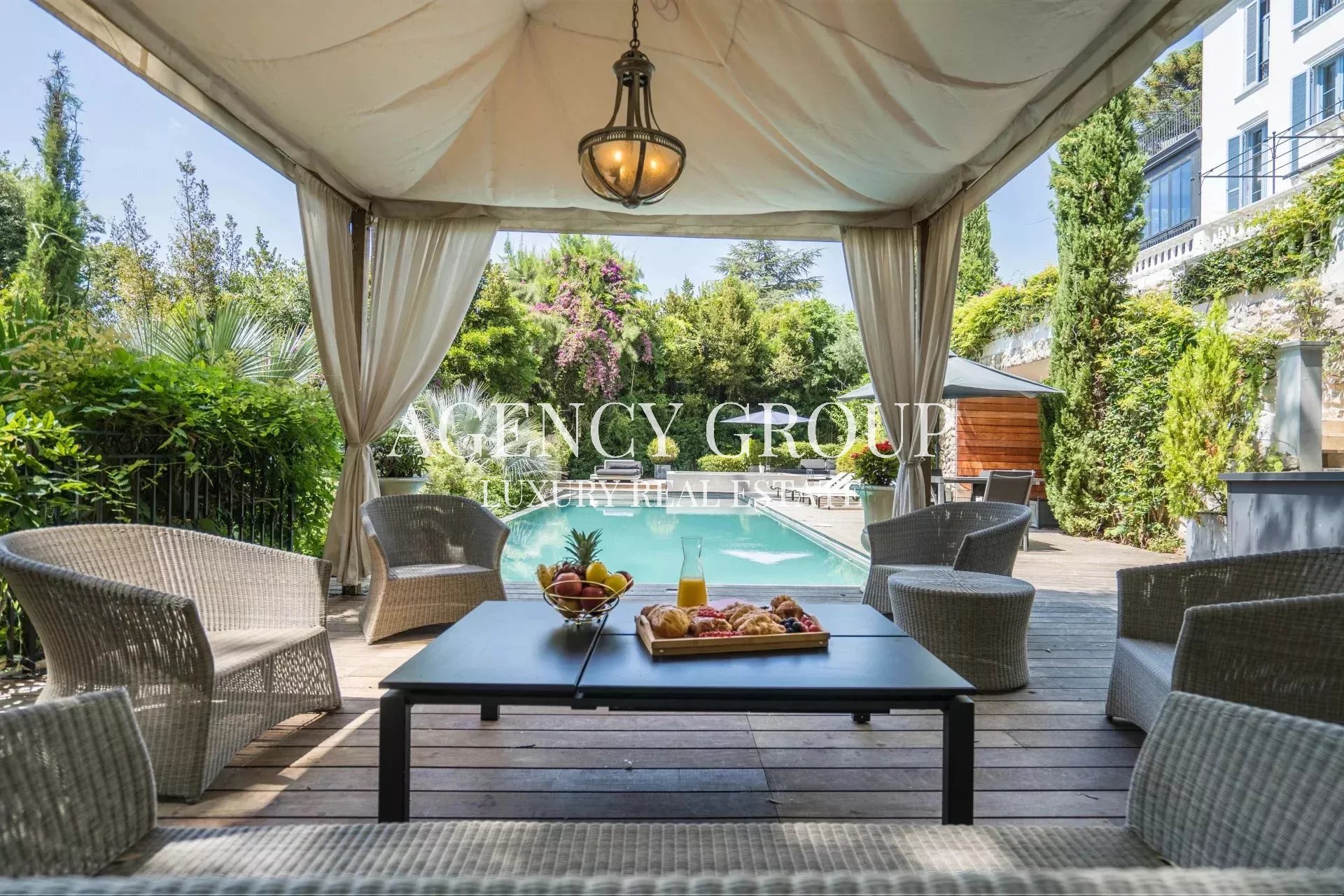 Outdoor cabana-style seating area with cream curtains, a central coffee table, and a view of a rectangular pool surrounded by greenery.