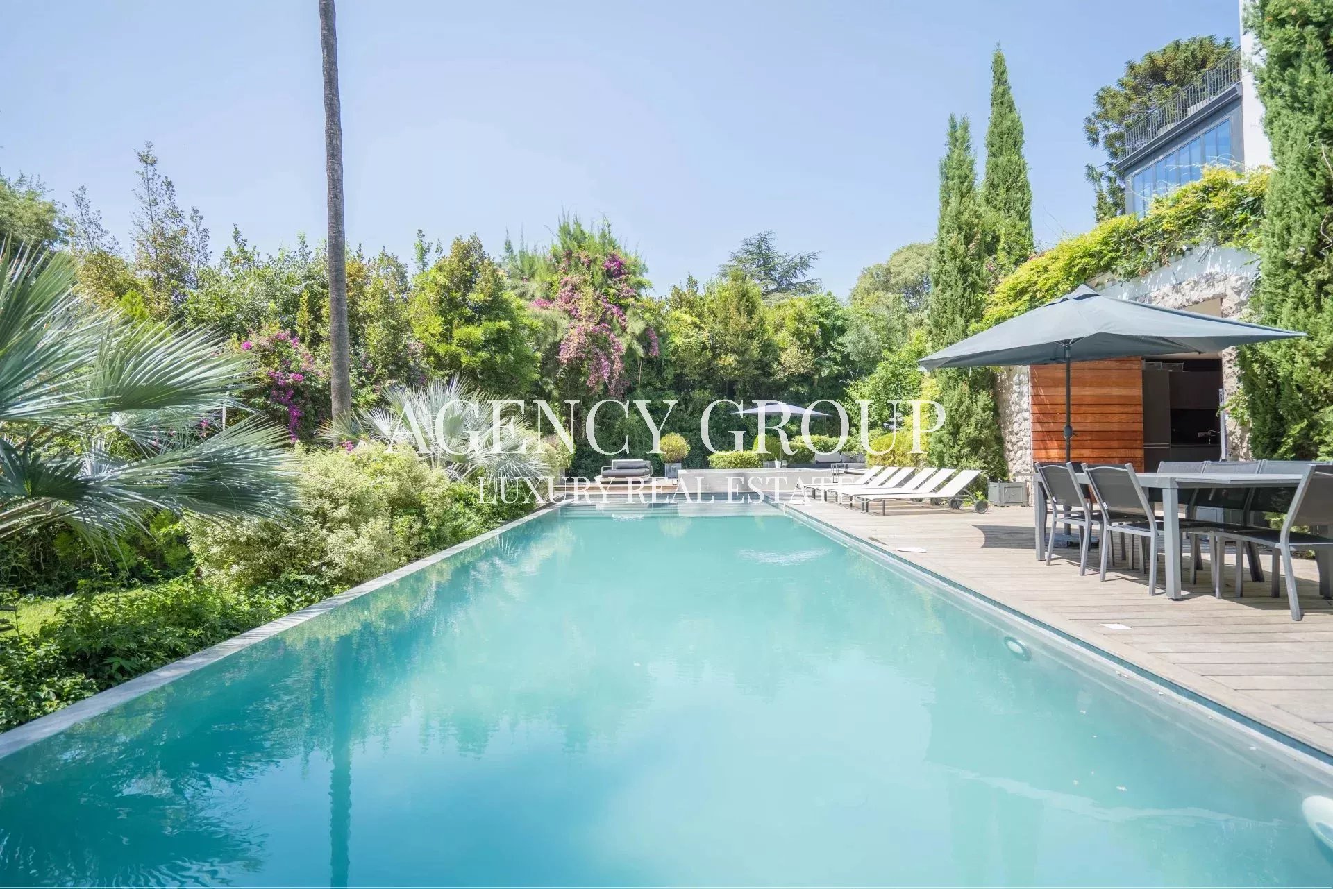 Poolside luxury scene with a rectangular pool, wooden deck, lounge chairs, and a dining table under a large umbrella; watermark reads 'Agency Group Luxury Real Estate'.