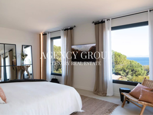 Bright bedroom with white bedding, neutral curtains, and large ocean-view windows; a wicker chair sits by the window.
