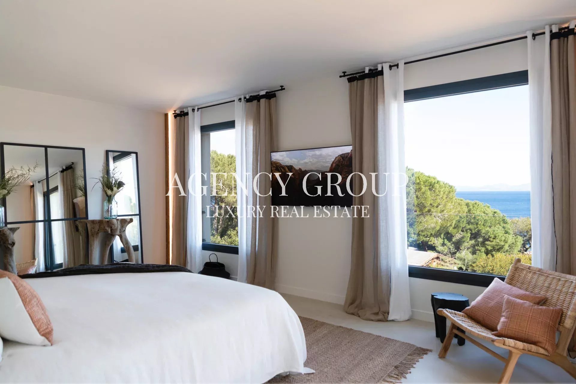 Bright bedroom with white bedding, neutral curtains, and large ocean-view windows; a wicker chair sits by the window.
