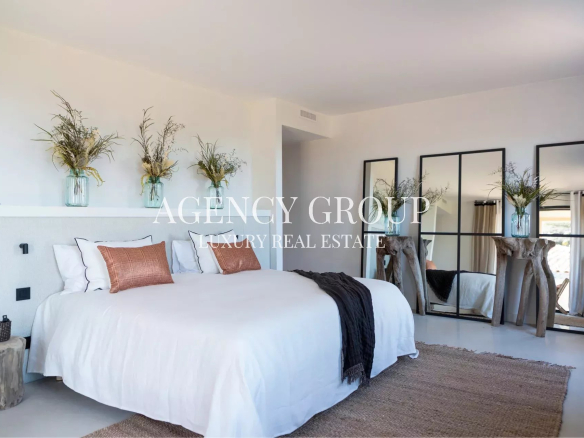 Bright luxury bedroom with a white bed, orange pillows, a dark throw, and tall greenery in glass jars on a shelf; real estate branding overlay reads ‘AGENCY GROUP LUXURY REAL ESTATE’.