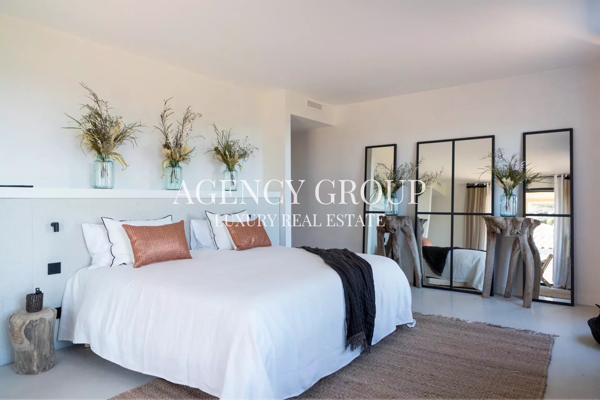 Bright luxury bedroom with a white bed, orange pillows, a dark throw, and tall greenery in glass jars on a shelf; real estate branding overlay reads ‘AGENCY GROUP LUXURY REAL ESTATE’.