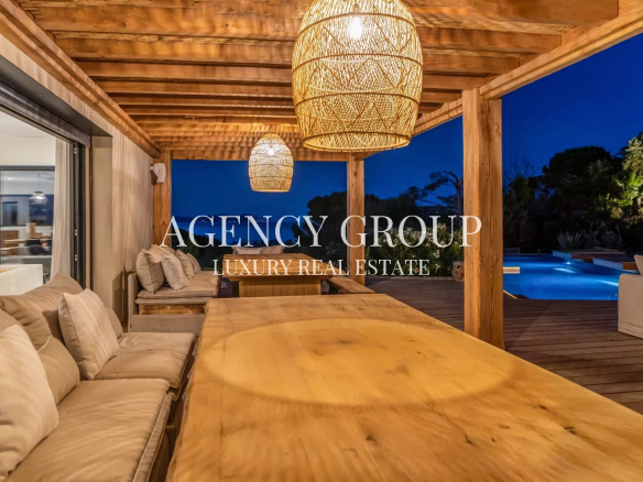 Luxurious outdoor patio at dusk with a long wooden dining table, beige cushioned seating, and woven pendant lights, overlooking a illuminated pool.