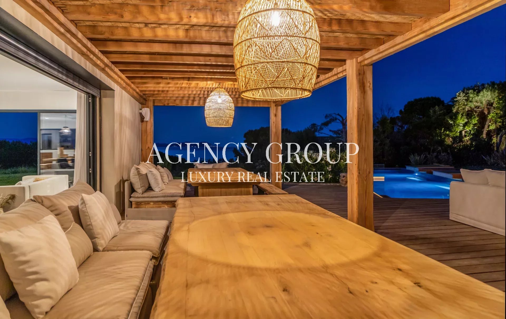 Luxurious outdoor patio at dusk with a long wooden dining table, beige cushioned seating, and woven pendant lights, overlooking a illuminated pool.