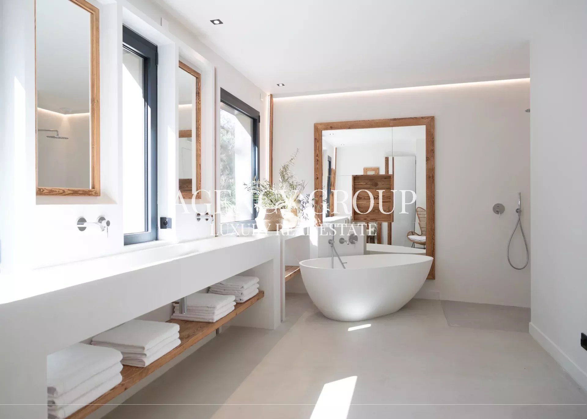 Bright minimalist bathroom with a freestanding oval tub, wooden-framed mirrors, and open-shelf towel storage.