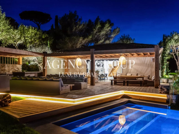 Luxurious outdoor lounge under a wooden pergola at night, with warm lighting and a blue-lit rectangular pool nearby.