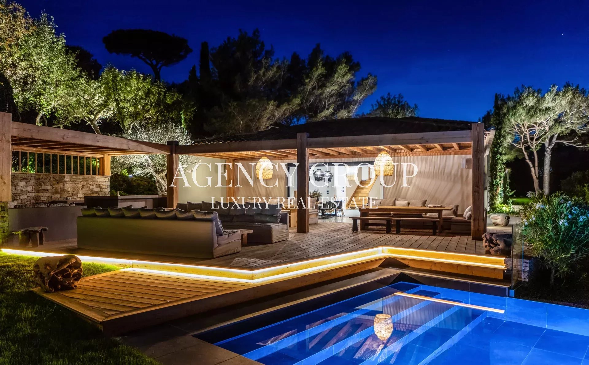 Luxurious outdoor lounge under a wooden pergola at night, with warm lighting and a blue-lit rectangular pool nearby.