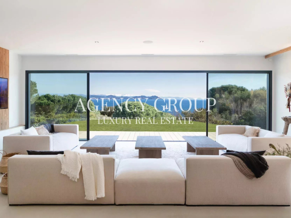 Bright modern living room with large glass doors opening to a lawn and ocean view; overlay reads 'AGENCY GROUP LUXURY REAL ESTATE'.