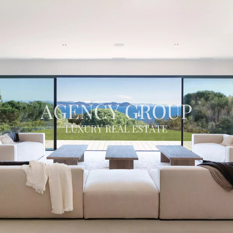 Bright modern living room with large glass doors opening to a lawn and ocean view; overlay reads 'AGENCY GROUP LUXURY REAL ESTATE'.
