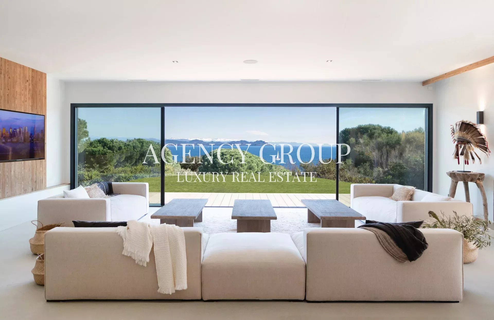 Bright modern living room with large glass doors opening to a lawn and ocean view; overlay reads 'AGENCY GROUP LUXURY REAL ESTATE'.