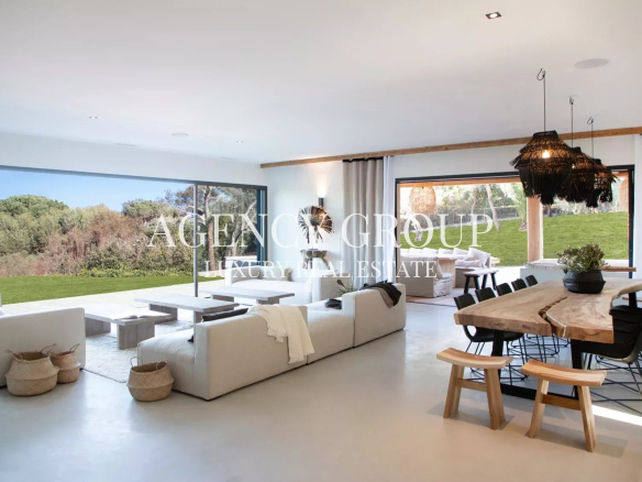 Open-plan luxury living room with white sectional sofas, a natural wood dining table, and floor-to-ceiling glass walls overlooking a green lawn and the ocean outside.