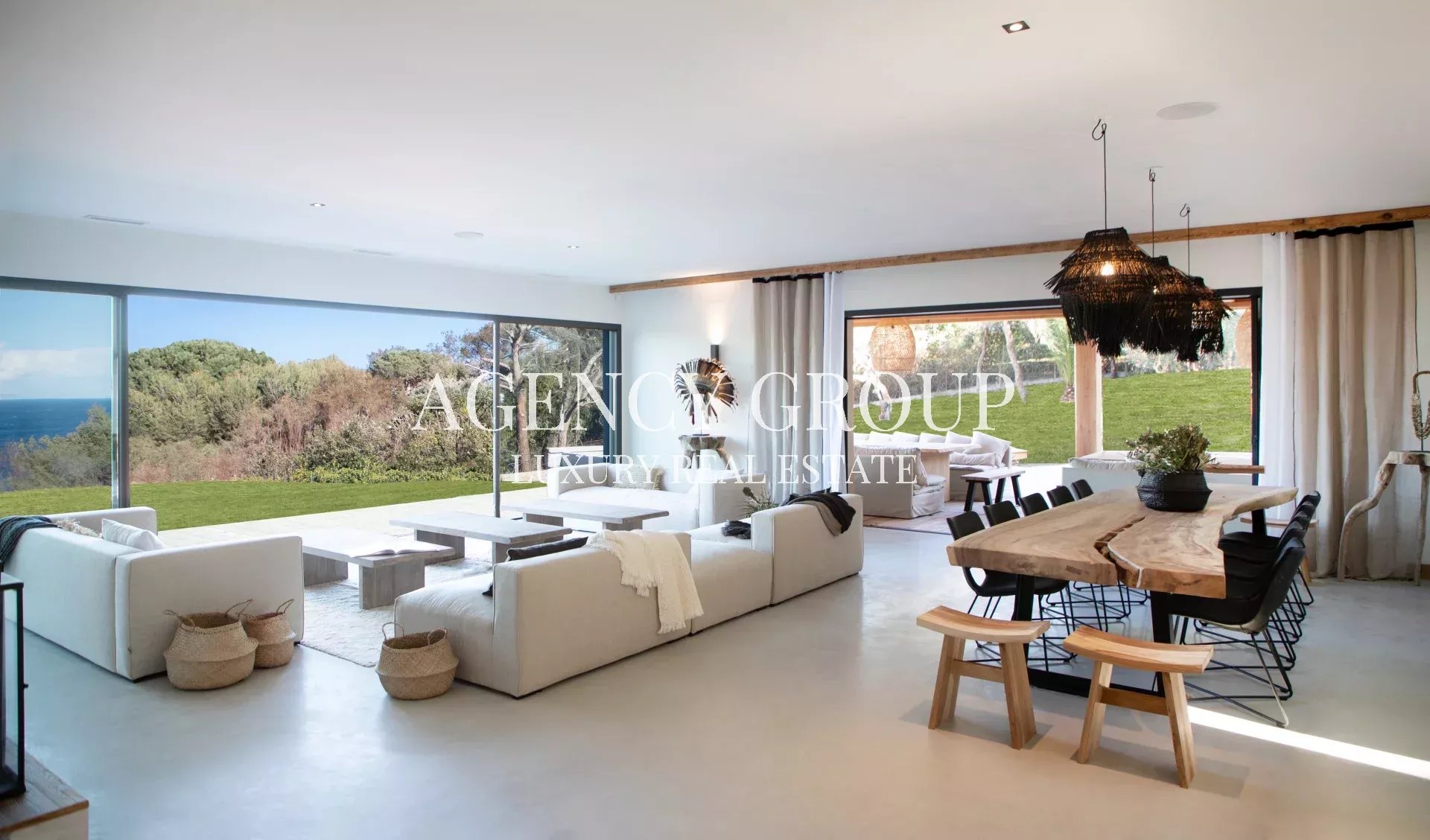 Open-plan luxury living room with white sectional sofas, a natural wood dining table, and floor-to-ceiling glass walls overlooking a green lawn and the ocean outside.