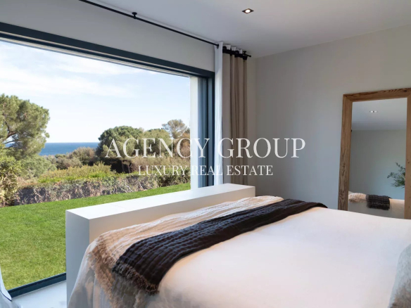 Bright bedroom with a large window revealing a green lawn, trees, and the ocean beyond; bed with white linens and a dark brown throw Blanket is across the room.