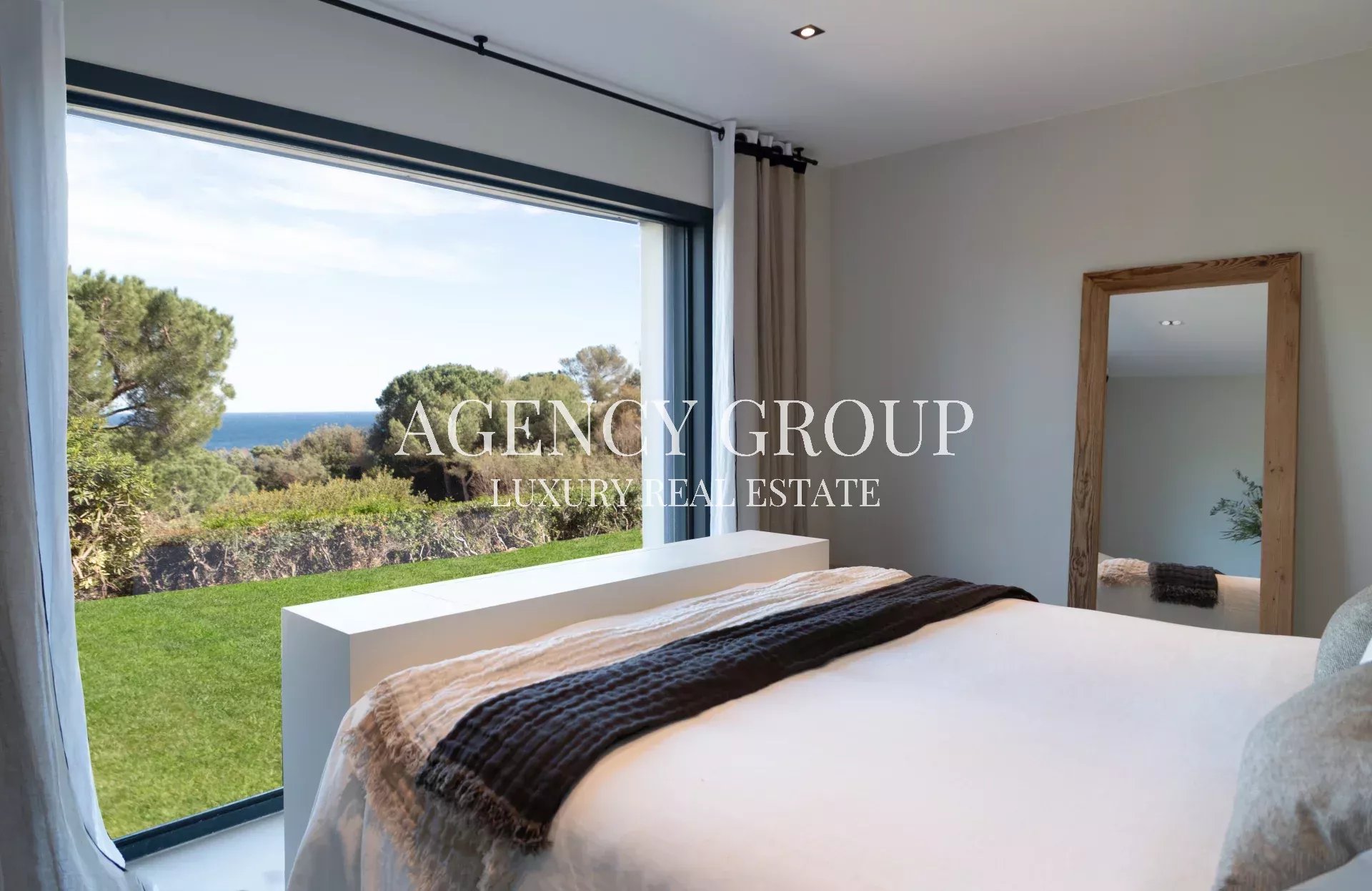 Bright bedroom with a large window revealing a green lawn, trees, and the ocean beyond; bed with white linens and a dark brown throw Blanket is across the room.