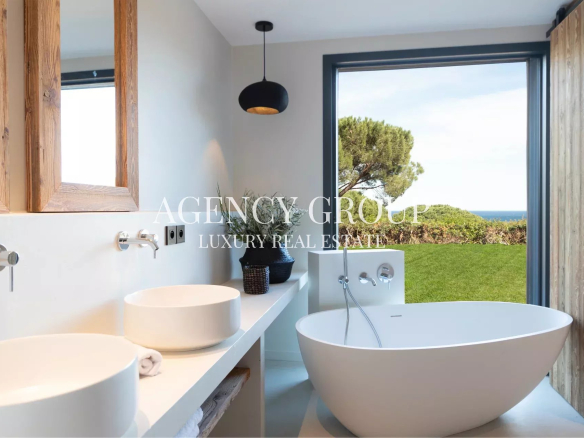 Bright modern bathroom with a white freestanding tub, double sinks, and wooden-framed mirrors, overlooking a lawn and the sea.