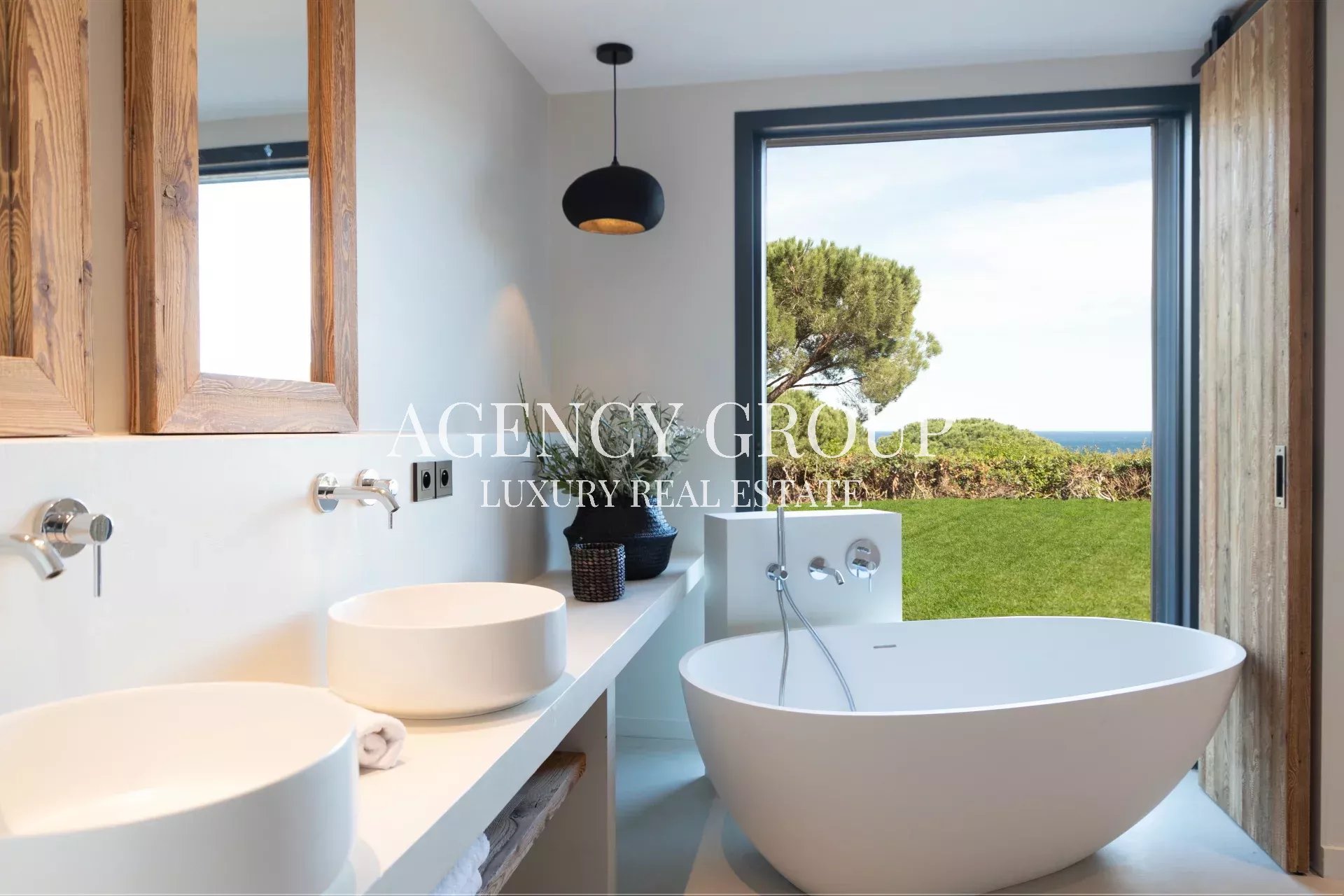 Bright modern bathroom with a white freestanding tub, double sinks, and wooden-framed mirrors, overlooking a lawn and the sea.