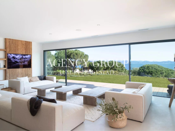 Luxurious living room with white sofas, wood panel accent wall, and floor-to-ceiling windows opening to a green lawn and ocean view.