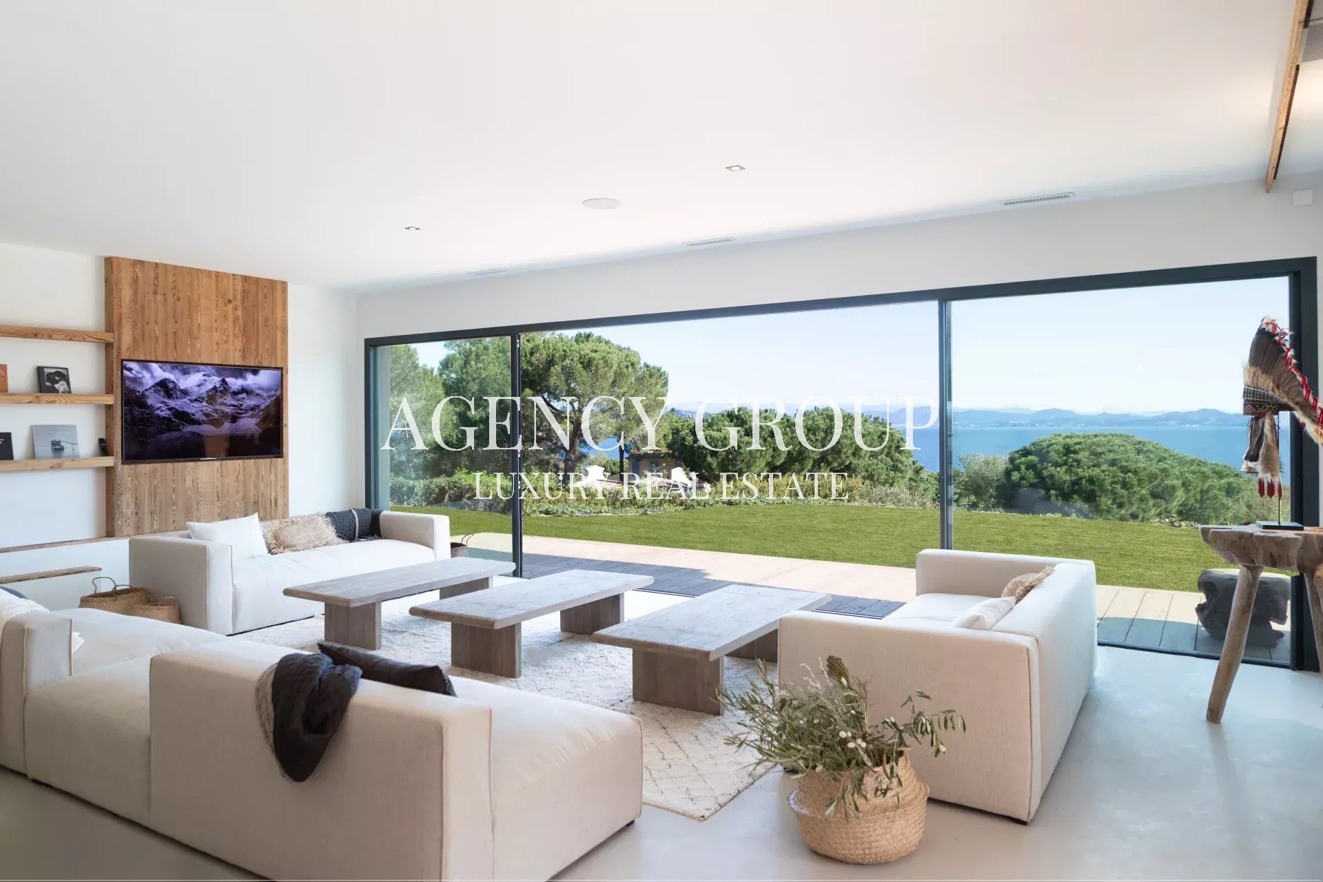 Luxurious living room with white sofas, wood panel accent wall, and floor-to-ceiling windows opening to a green lawn and ocean view.