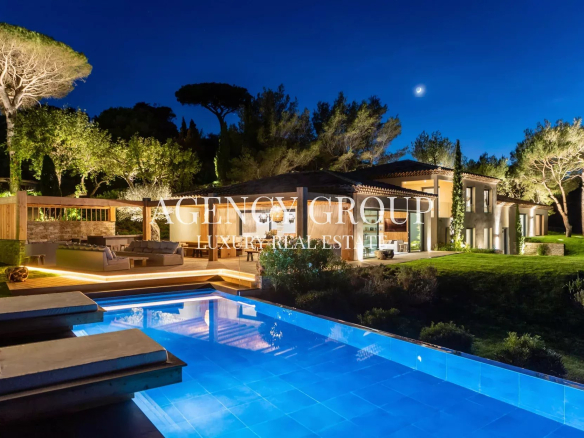 Luxurious nighttime backyard with a modern villa, warm terrace lighting, and a blue rectangular pool. Eventually a crescent moon in the sky.