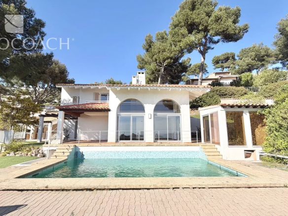 White villa with arched windows facing a rectangular pool on a sunny day; lush trees in the background.
