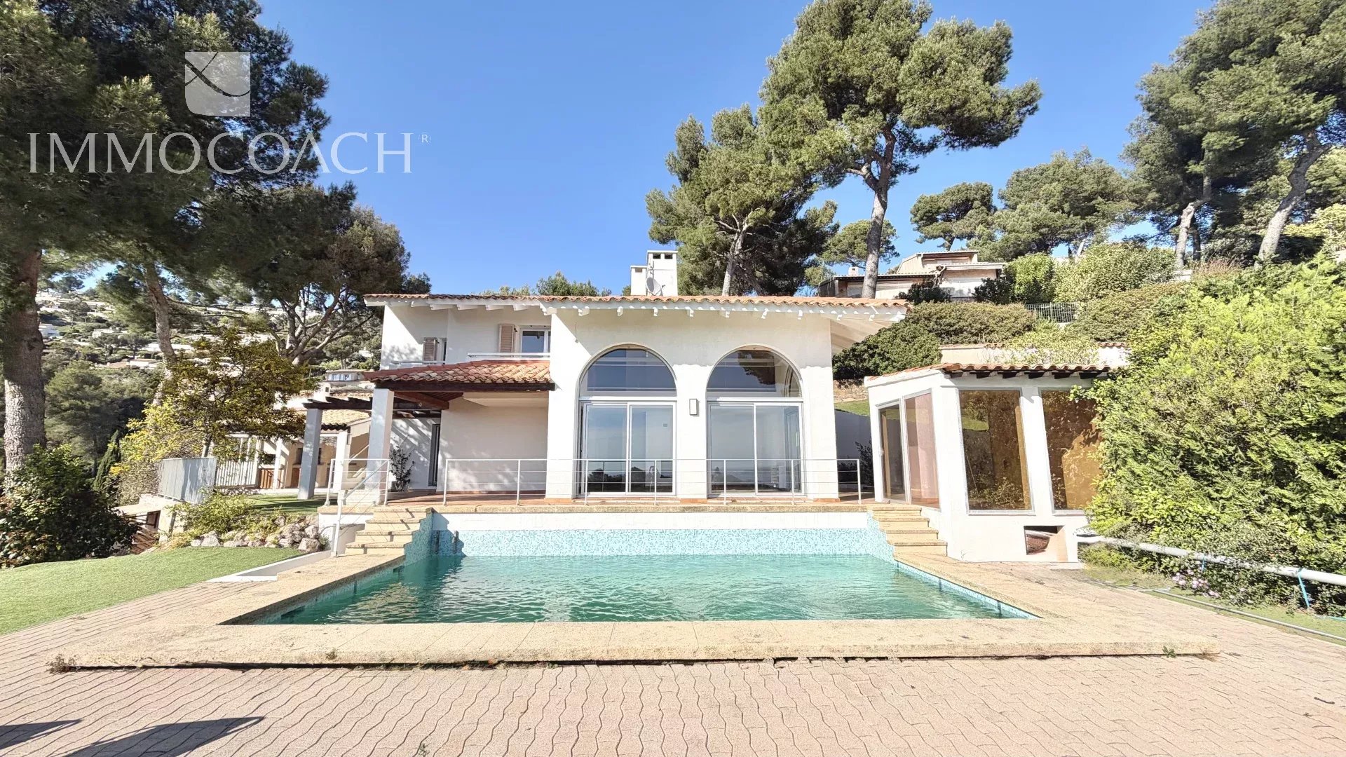 White villa with arched windows facing a rectangular pool on a sunny day; lush trees in the background.
