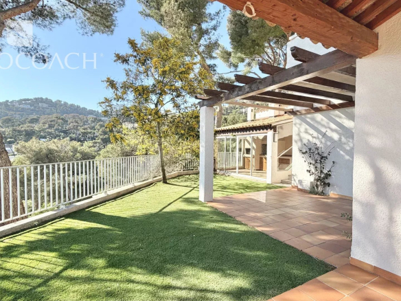 Terrace with a grassy yard, white railing, and a wooden pergola overlooking a hillside landscape on a sunny day.