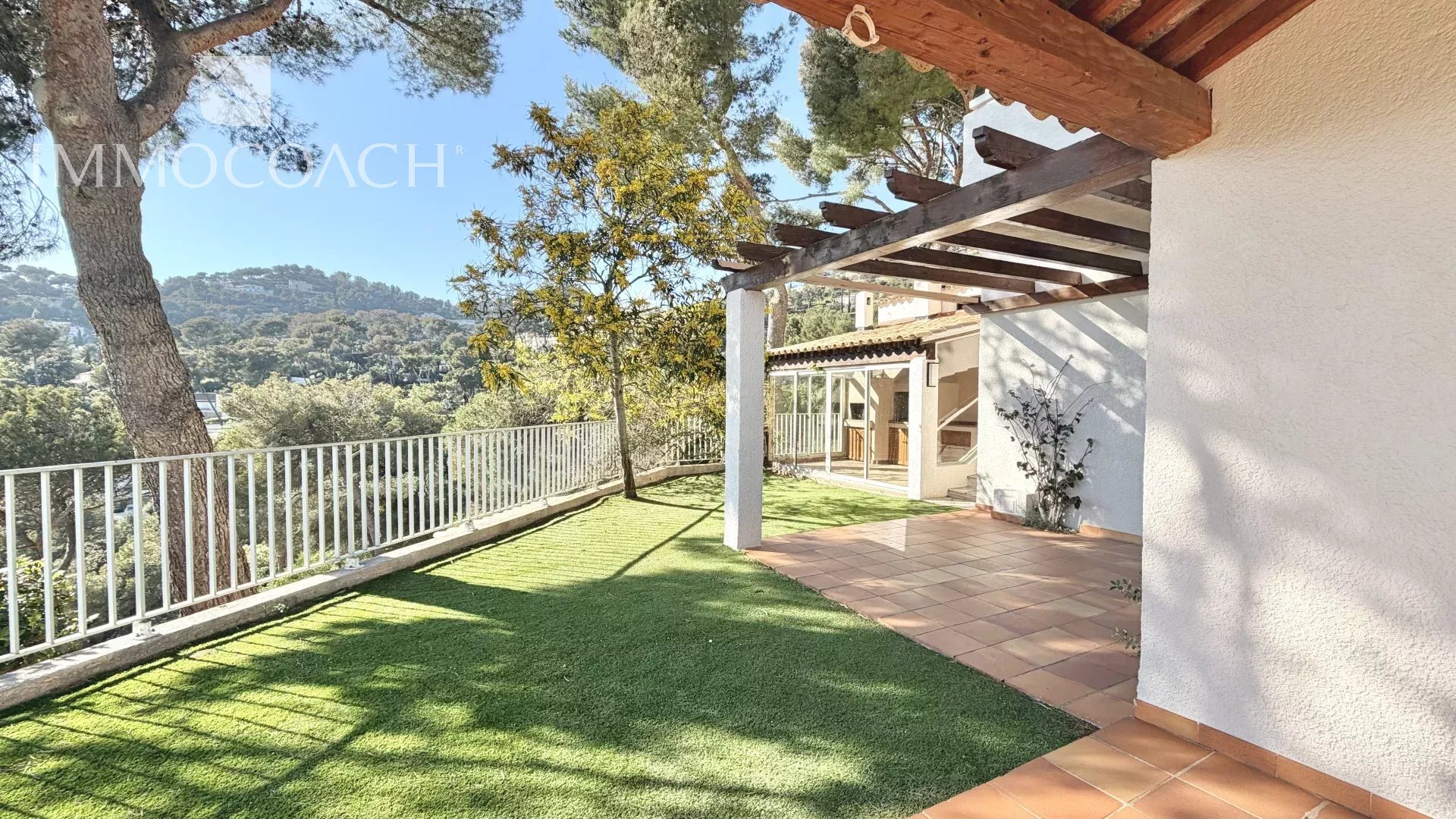 Terrace with a grassy yard, white railing, and a wooden pergola overlooking a hillside landscape on a sunny day.