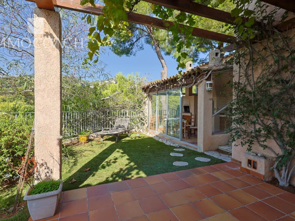 Terracotta-tiled patio opens to a sunlit garden with a lounge chair, glass sliding doors to the house, and a vine-covered pergola overhead.
