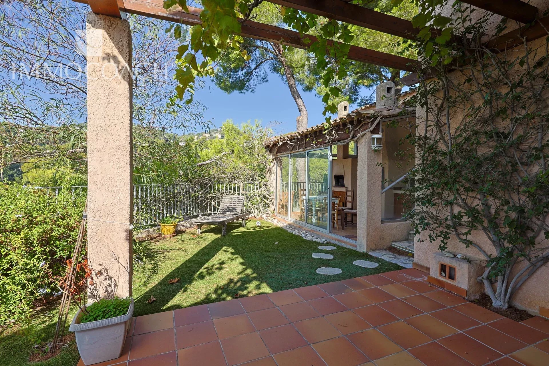 Terracotta-tiled patio opens to a sunlit garden with a lounge chair, glass sliding doors to the house, and a vine-covered pergola overhead.