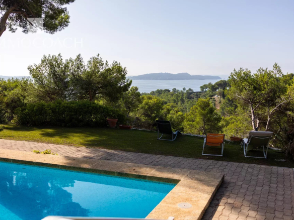 Poolside scene with four lounge chairs on a paved patio, overlooking a lush garden and the blue sea.