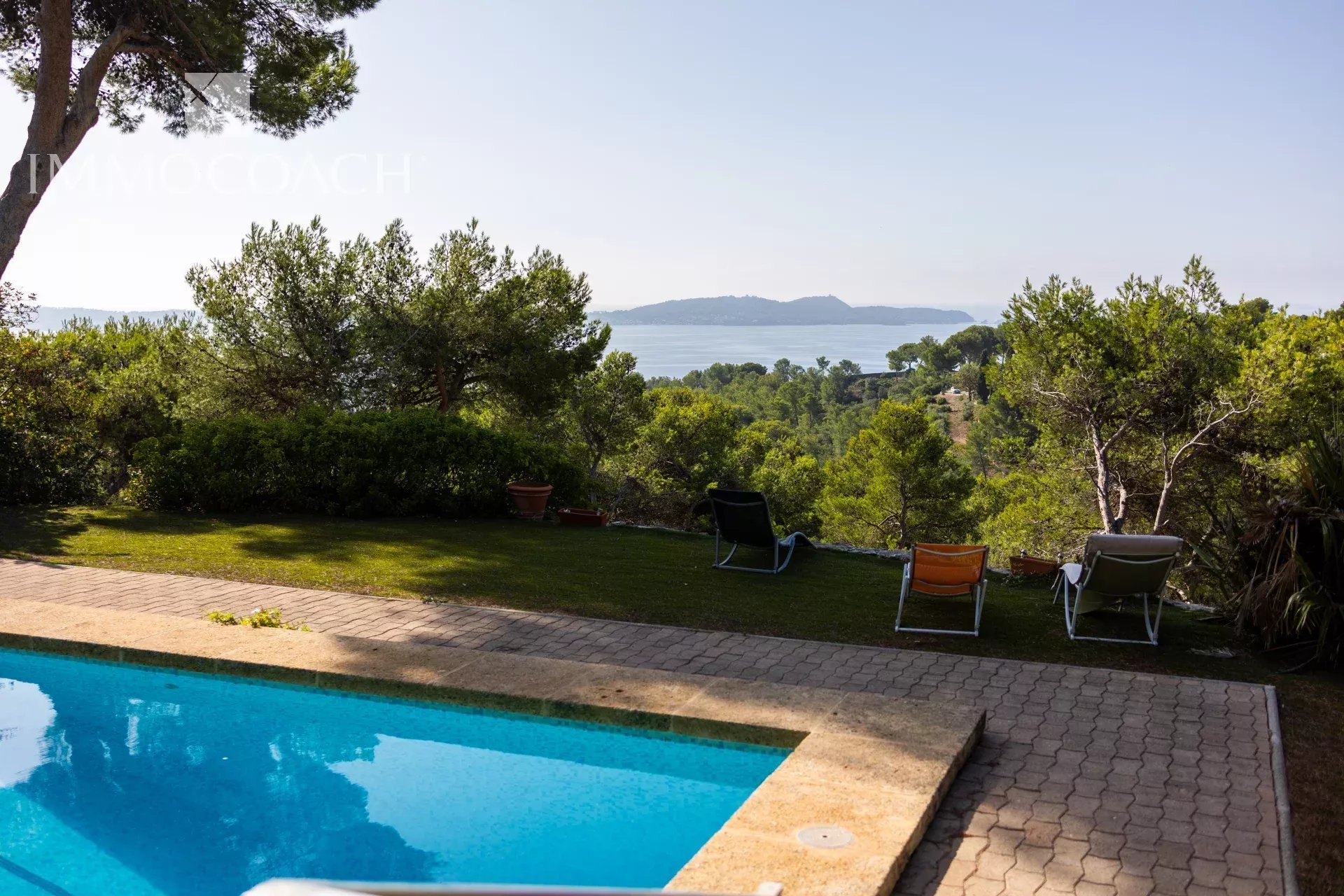 Poolside scene with four lounge chairs on a paved patio, overlooking a lush garden and the blue sea.