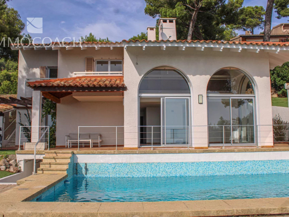 Modern white villa with a terracotta roof, large arched windows, and a blue pool in the foreground. Has a stone deck and metal railing.