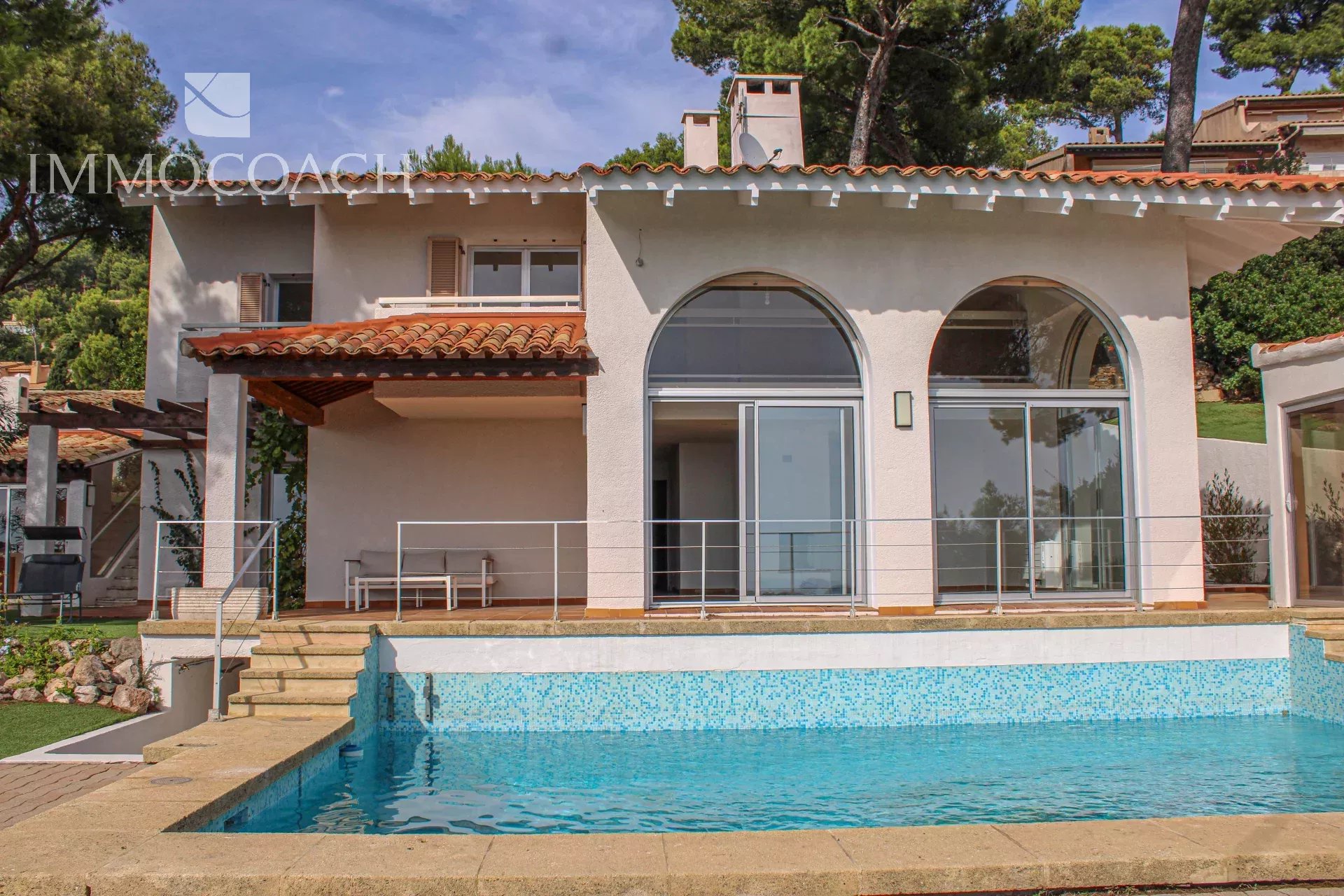 Modern white villa with a terracotta roof, large arched windows, and a blue pool in the foreground. Has a stone deck and metal railing.