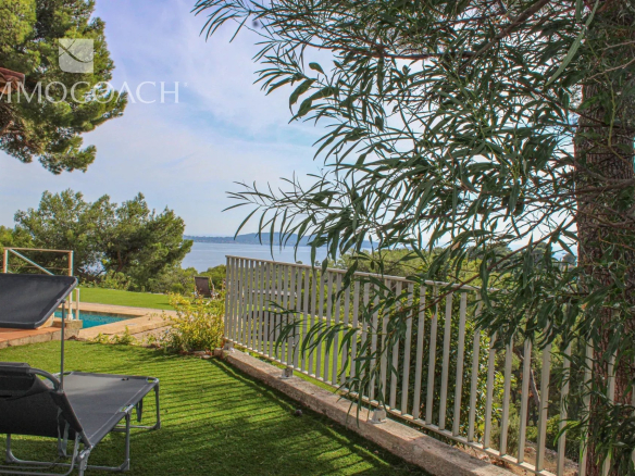 Backyard with two black lounge chairs on a green lawn, a white railing, and a sea view beyond trees under a blue sky, watermark in top-left.