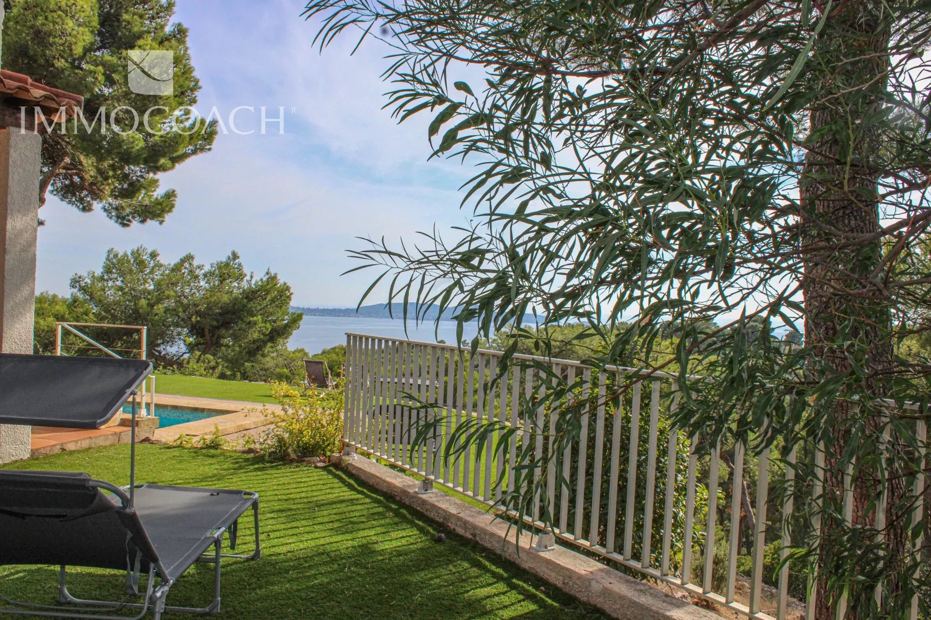Backyard with two black lounge chairs on a green lawn, a white railing, and a sea view beyond trees under a blue sky, watermark in top-left.