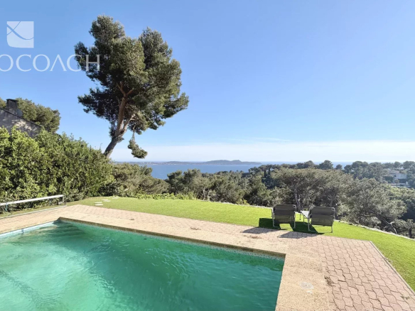 Pool with turquoise water overlooking a sea view, lounge chairs on a sunny patio, blue sky and greenery surrounding. (Immocoach watermark)