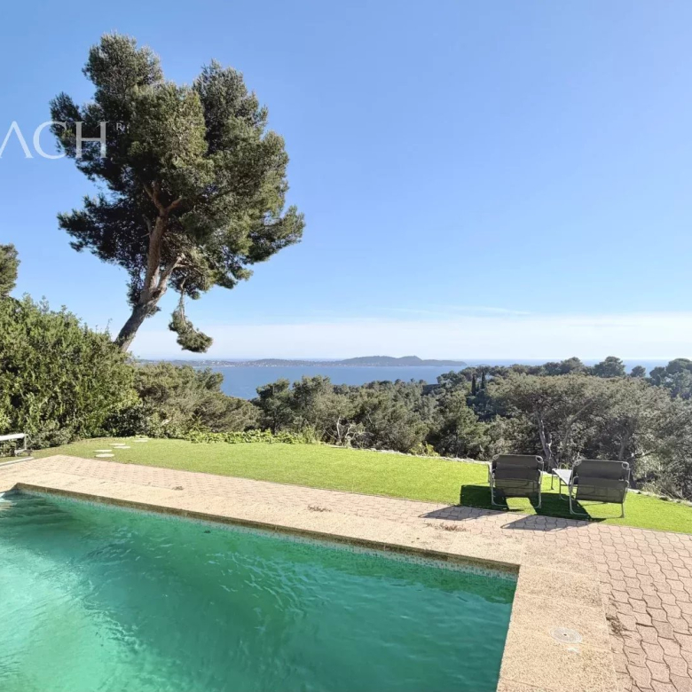 Pool with turquoise water overlooking a sea view, lounge chairs on a sunny patio, blue sky and greenery surrounding. (Immocoach watermark)