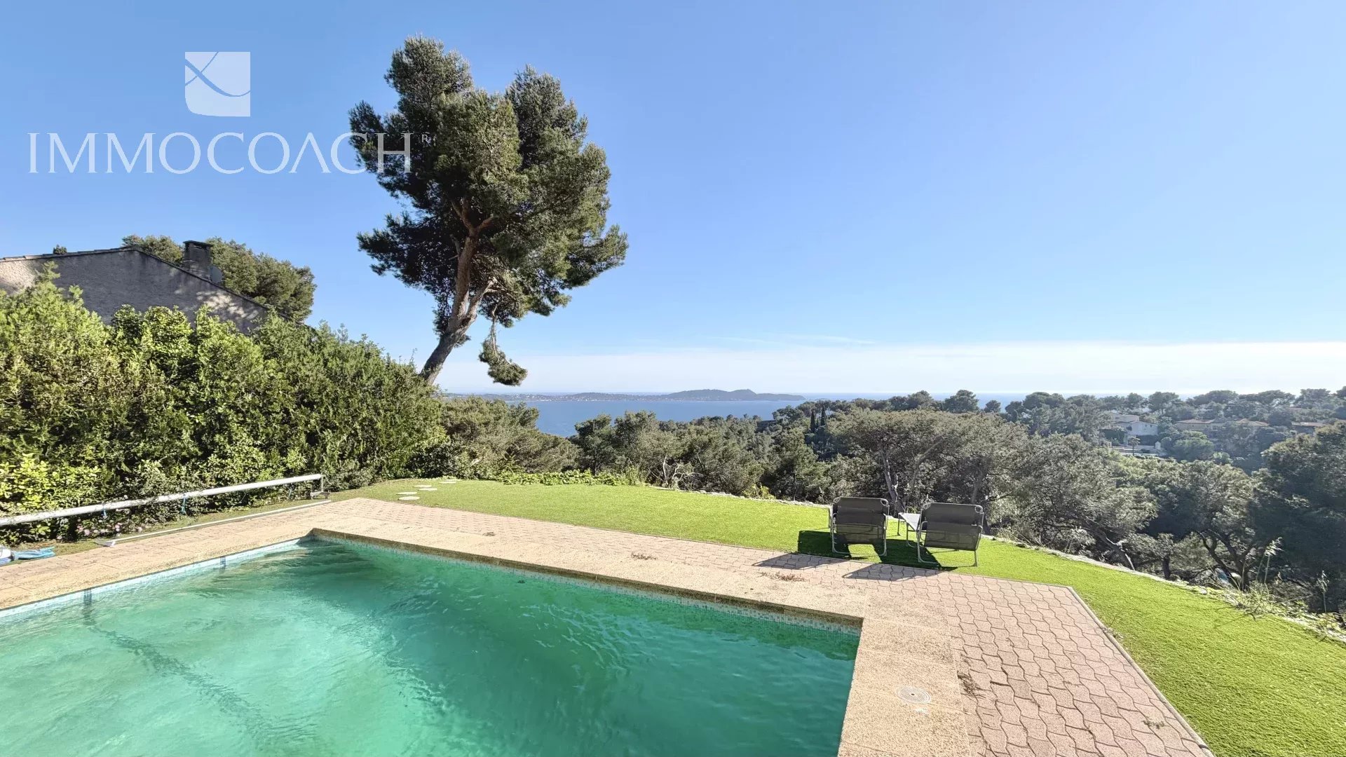 Pool with turquoise water overlooking a sea view, lounge chairs on a sunny patio, blue sky and greenery surrounding. (Immocoach watermark)