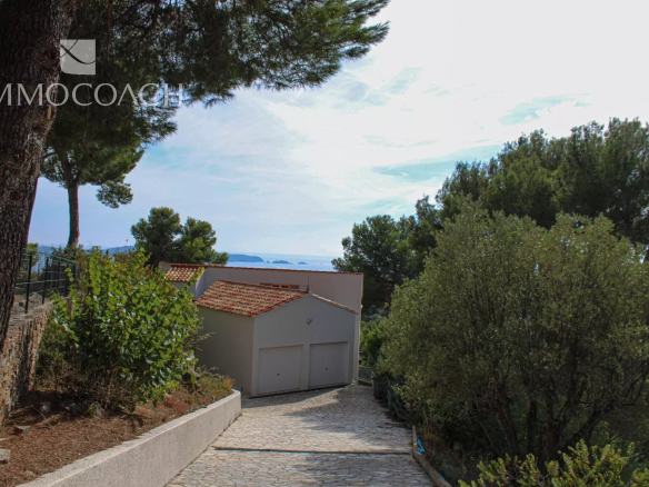 Stone driveway leads to a white, two-car garage with a red-tiled roof, flanked by trees and shrubs with a distant sea view.