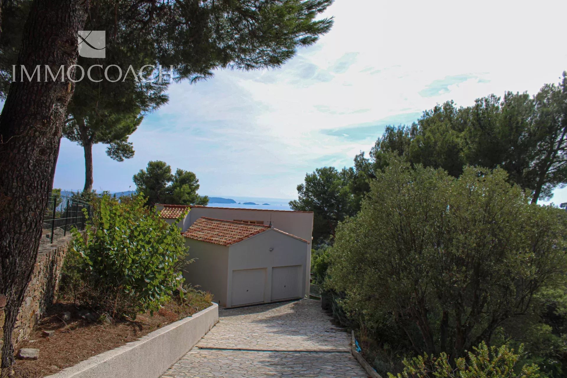 Stone driveway leads to a white, two-car garage with a red-tiled roof, flanked by trees and shrubs with a distant sea view.