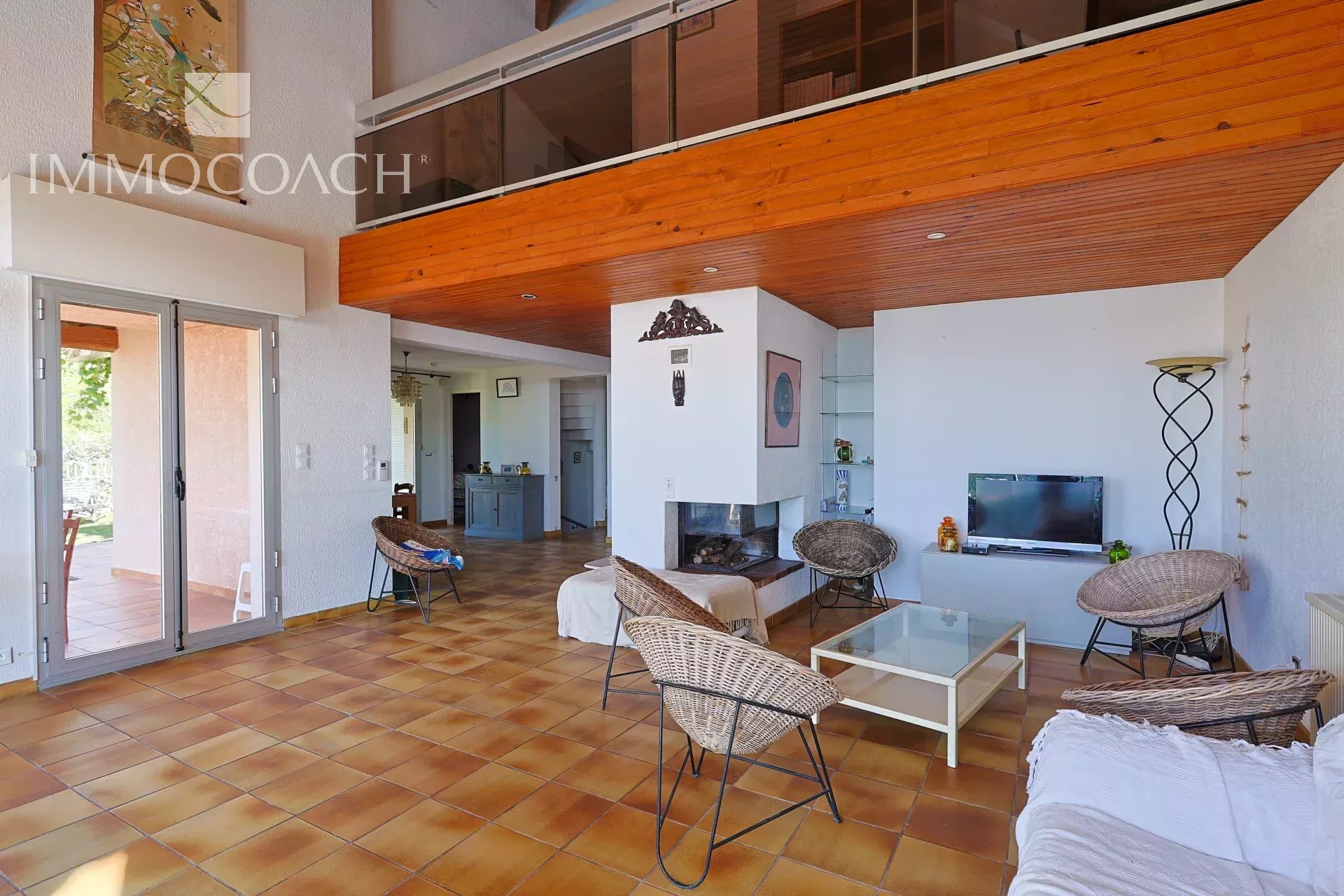 Spacious living room with terracotta tile floor, white walls, a central fireplace, and wicker chairs around a glass coffee table.